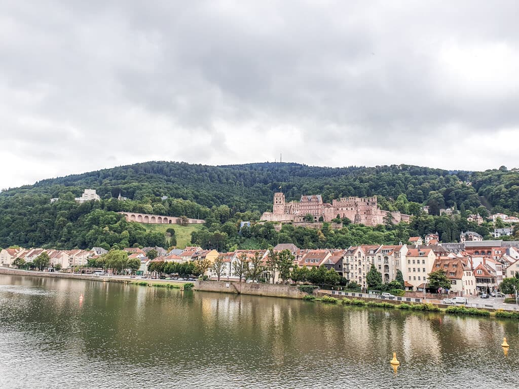 Blick über den Neckar auf eine Häuserreihe der Heidelberger Altstadt mit dem Schloss darüber und dem Königstuhl