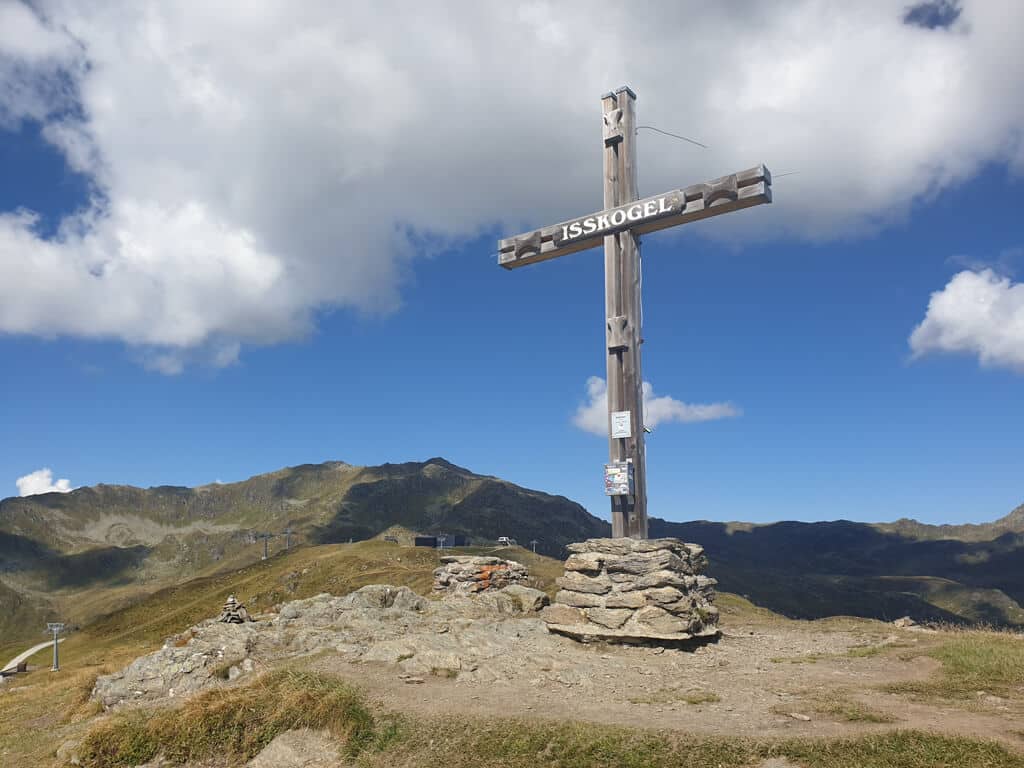 Gipfelkreuz aus Holz "Isskogel" - rundherum grüne Berggipfel und linksseitig einige Stützen eines Sessellift