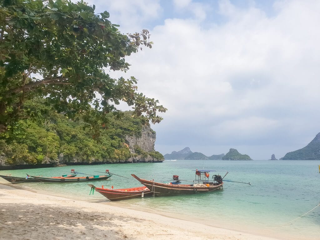 Longtailboote im flachen Wasser, daneben ein Sandstrand und im Hintergrund kleine Insel mit Felsen