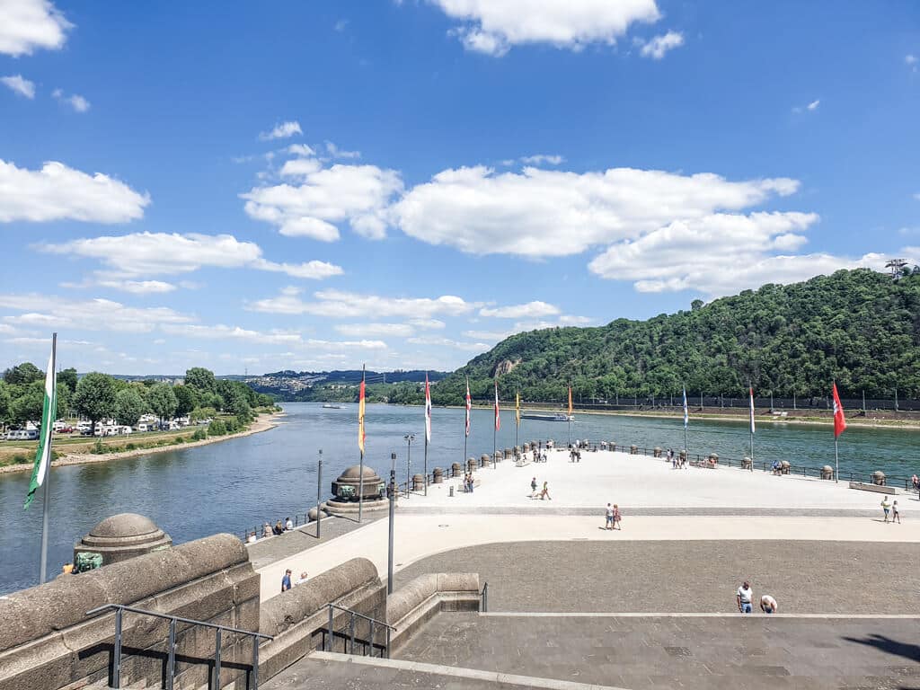 Blick über einen großen Platz mit Länderflaggen auf den Zusammenfluss von Rhein und Mosel am Deutschen Eck