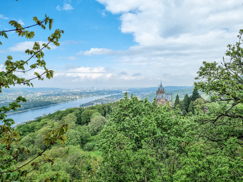 Blick über grüne Bäume auf das Schloss Drachenburg; links darunter fließt der Rhein. Zu beiden Seiten des Rheins sind Städte zu sehen.