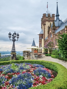 Blick über ein buntes Blumenbeet und einen Weg, der links am Schloss Drachenburg entlangführt.