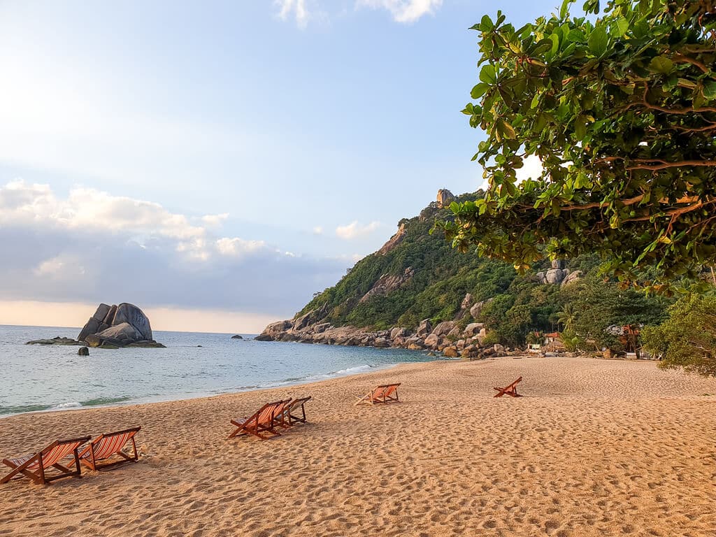 Liegestühle am Sandstrand, dahinter ein grüner Hügel und das Meer