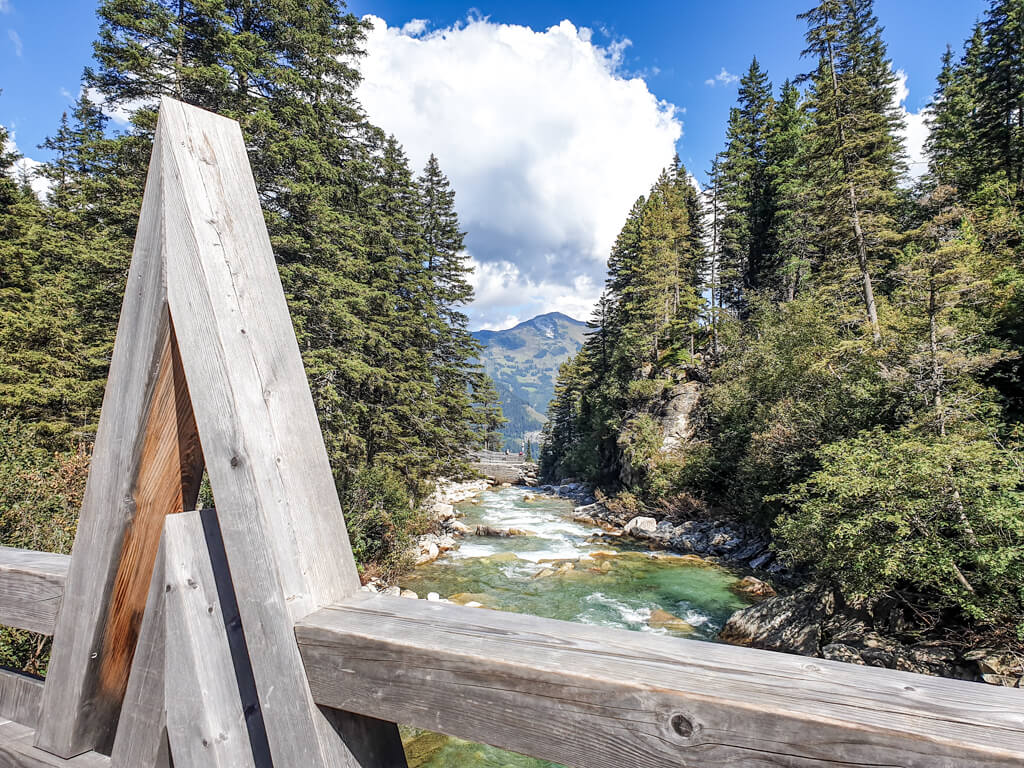 Blick über das Holzgeländer eine Brücke auf einen kleinen Fluss mit einigen Steinen und grünlichem Wasser. Der Fluss fließt zwischen Bäumen und Bergen entlang