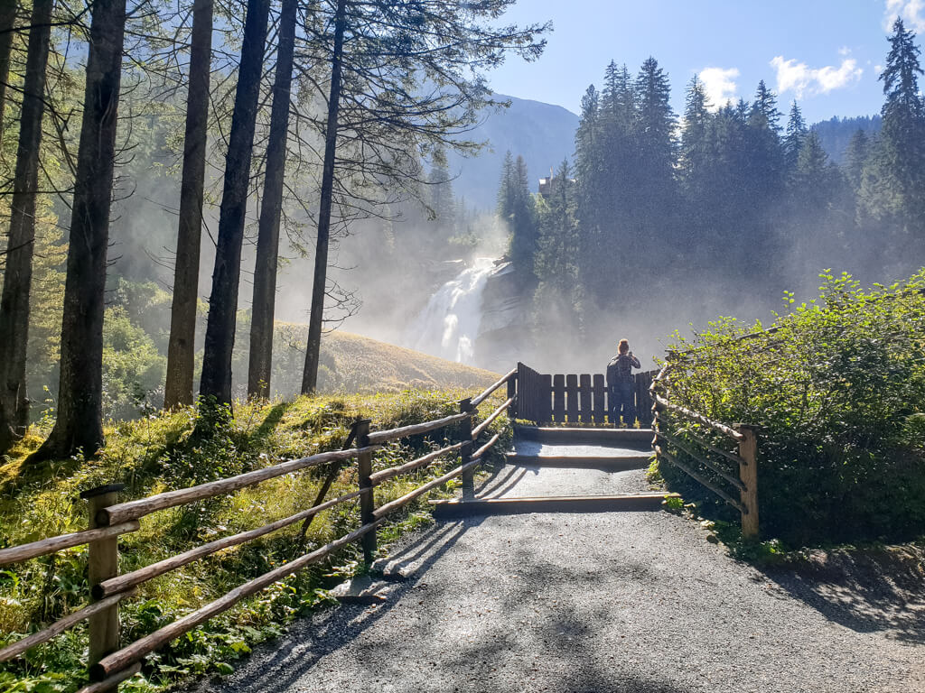 Weg führt zu einer Aussichtskanzel, die mit einem Holzzaun gesichert ist. Dahinter ist ein hoher Wasserfall sowie Bäume zu sehen.