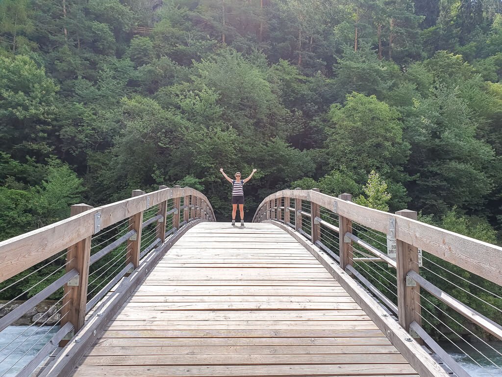 Frau steht mitten auf einer gebogenen Holzbrücke über einen Fluss