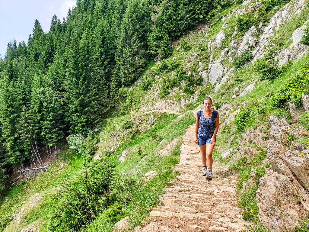 Frau steht auf einem schmalen Bergweg, im Hintergrund Wald, links ein abfallender Hang mit Wiese