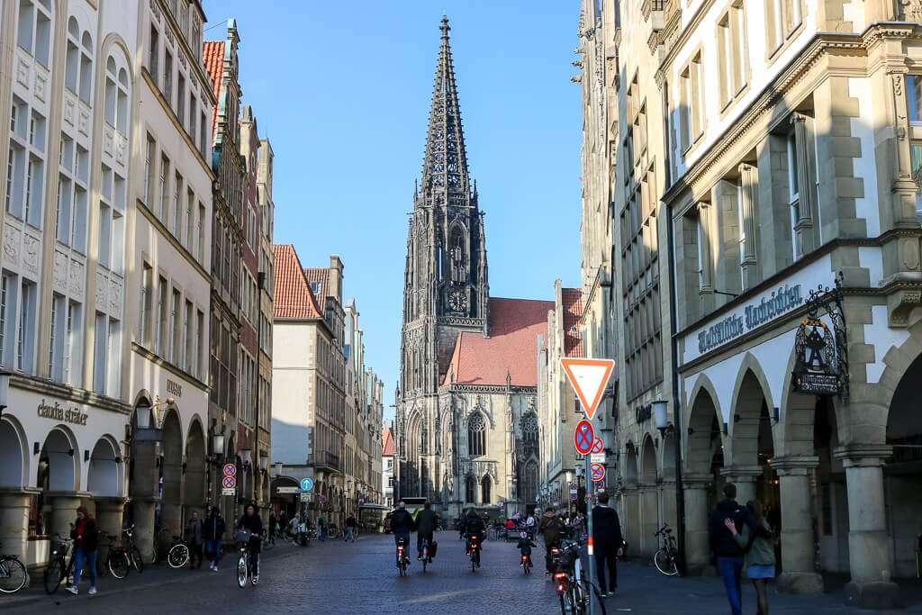 Prinzipalmarkt Münster mit Blick auf die Lambertikirche. Blick auf eine Kirche mit gotischem Kirchturm. Davor befinden sich in einer Flucht Giebelhäuser mit Bogengängen