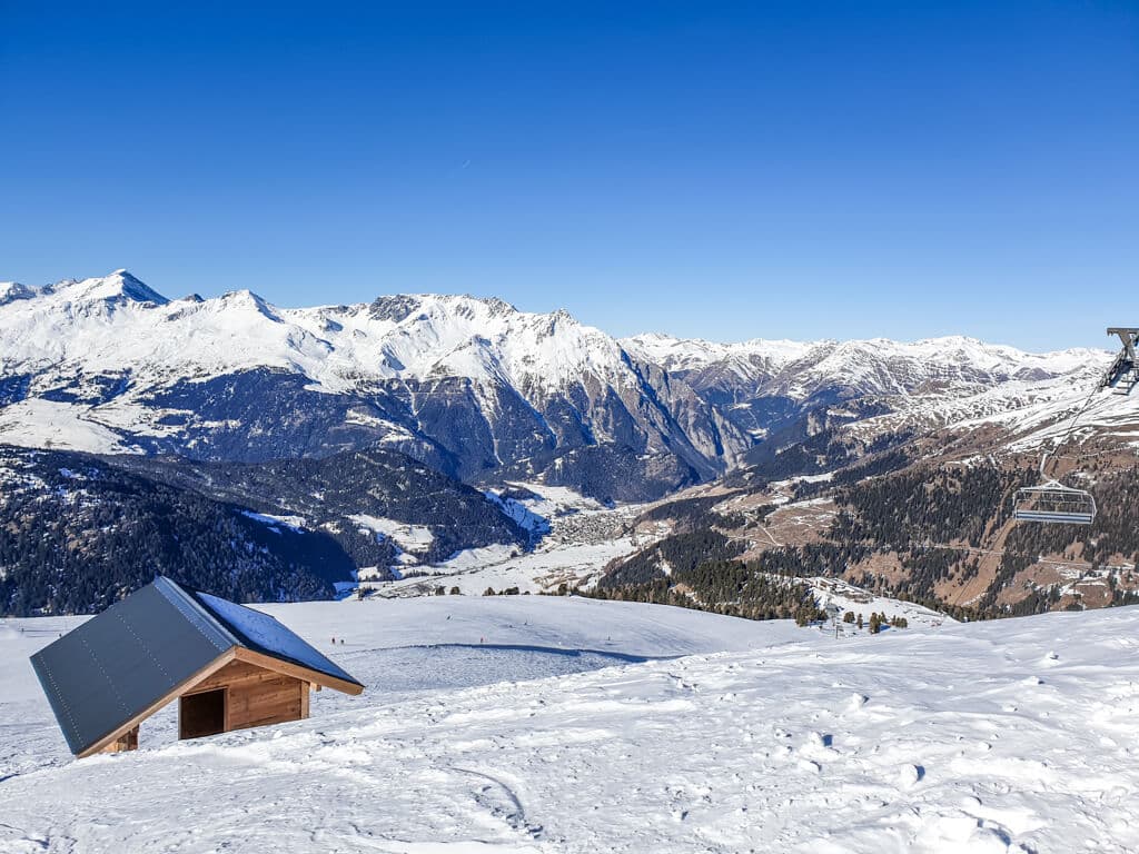 Blick auf ein Bergpanorama mit schneebedeckten Bergen - unten im Tal ist der Ort Nauders sichtbar. Oben am Berg steht eine Holzhütte im Schnee