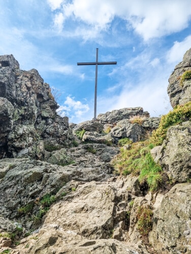 Ein großes Metallkreuz steht auf einem felsigen Hügel unter einem teilweise bewölkten blauen Himmel. Der schroffe Pfad, der zum Kreuz hinaufführt, ist von Felsbrocken und Grasflächen umgeben.
