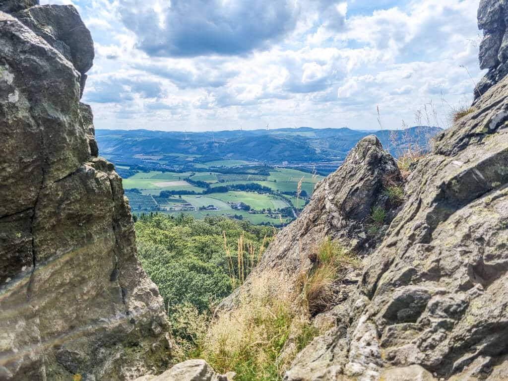 Eine malerische Aussicht auf ein Tal mit Flecken grüner Felder, Hügel und teilweise bewölktem Himmel, gesehen durch eine schmale Felsspalte. Im Vordergrund sind schroffe Felsformationen und ein paar wilde Grashalme zu sehen.
