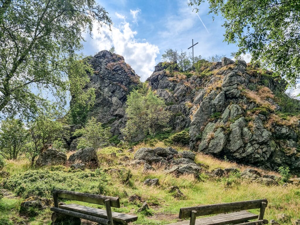 Ein felsiger Hügel mit einem Kreuz auf seiner Spitze ist von Grün umgeben und der Himmel ist teilweise bewölkt. Im Vordergrund stehen zwei Holzbänke, die einen Blick auf die üppige Landschaft bieten. Bäume und Sträucher sind in der Umgebung verstreut.