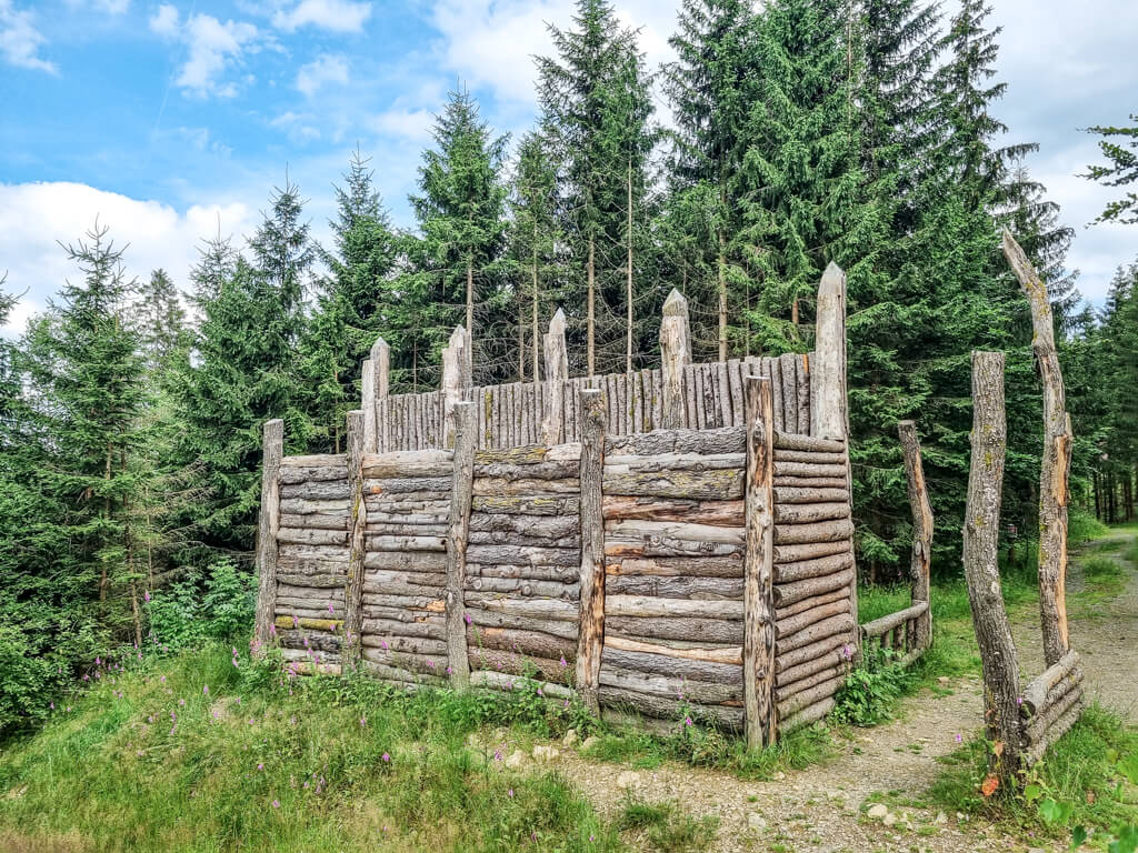 In einem Wald steht eine hölzerne, festungsähnliche Struktur, die aus Baumstämmen mit spitzen Spitzen gebaut wurde. Sie ist von hohen immergrünen Bäumen umgeben und in Tageslicht getaucht. Der Himmel ist teilweise bewölkt mit blauen Flecken. Ein Pfad führt hinauf zur Wall.
