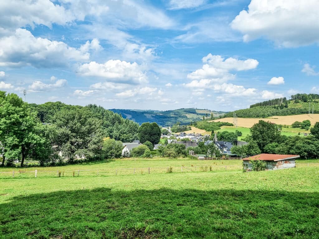 Ausblick über grüne Wiesen zu einer kleinen Ortschaft. Im Hintergrund erheben sich kleinere begrünte Hügel vor dem blauen, leicht bewölkten Himmel