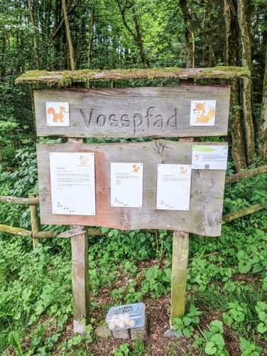 Holztafel im Wald mit Aufschrift "Vosspfad" und zwei Füchsen als Symbol daneben. Tafel ist eingebettet in grünes Laub am Rande eines Waldes