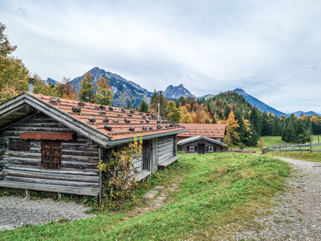 Holzhütten mit roten Ziegeldächern stehen in einer grasbewachsenen Landschaft. Herbstbäume und Berge bilden den Hintergrund unter einem bewölkten Himmel. Ein Schotterweg führt durch die Szene und verleiht eine rustikale, ruhige Atmosphäre.
