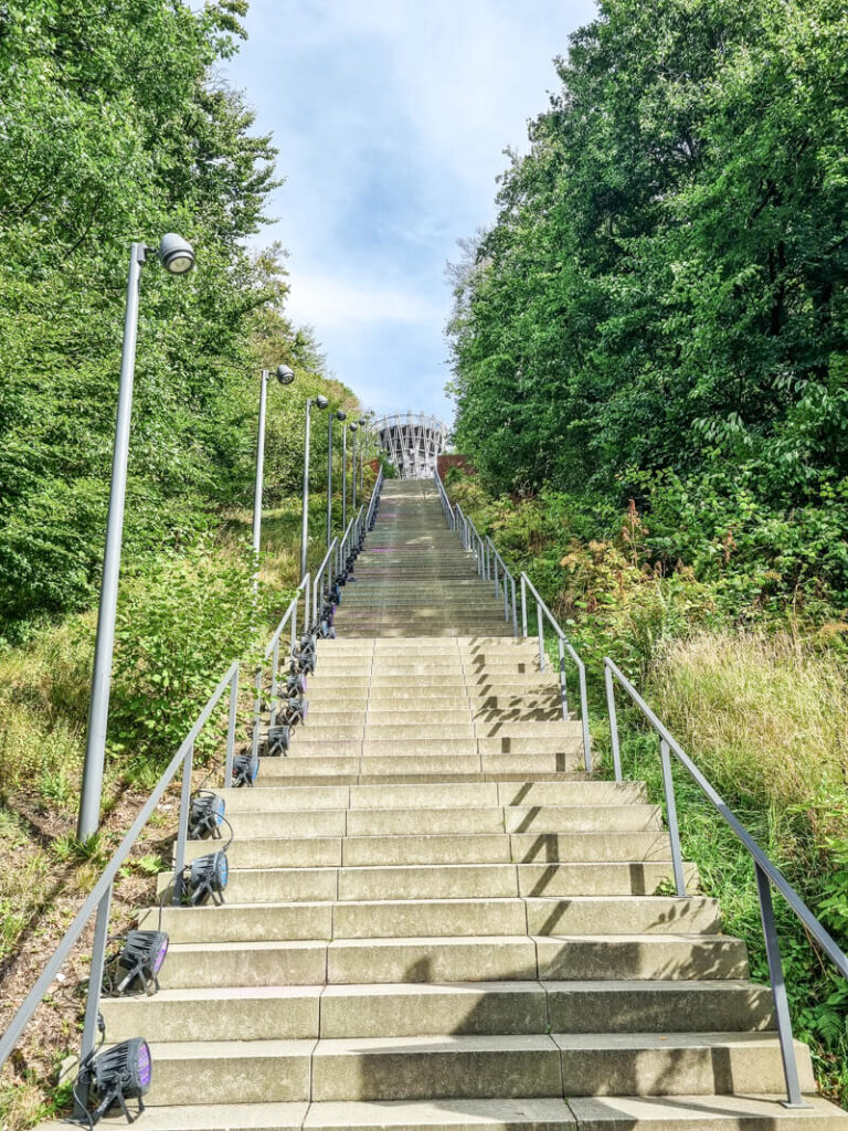 Eine lange, breite Außentreppe mit Metallgeländern und Lampen auf beiden Seiten führt durch grüne, belaubte Bäume hinauf zu einem Gebäude an der Spitze unter einem teilweise bewölkten Himmel.