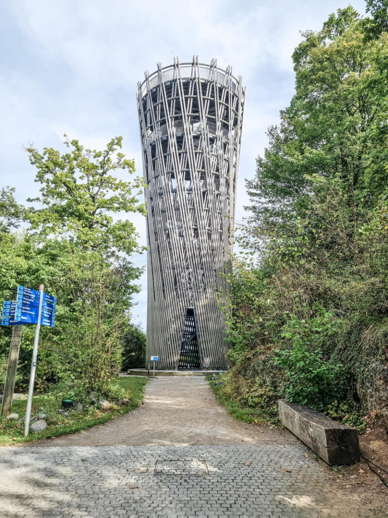 Ein hoher, zylindrischer Aussichtsturm mit einem sich kreuzenden Metallmuster steht zwischen grünen Bäumen entlang eines gepflasterten Weges, mit einer Holzbank und blauen Wegweisern in der Nähe.