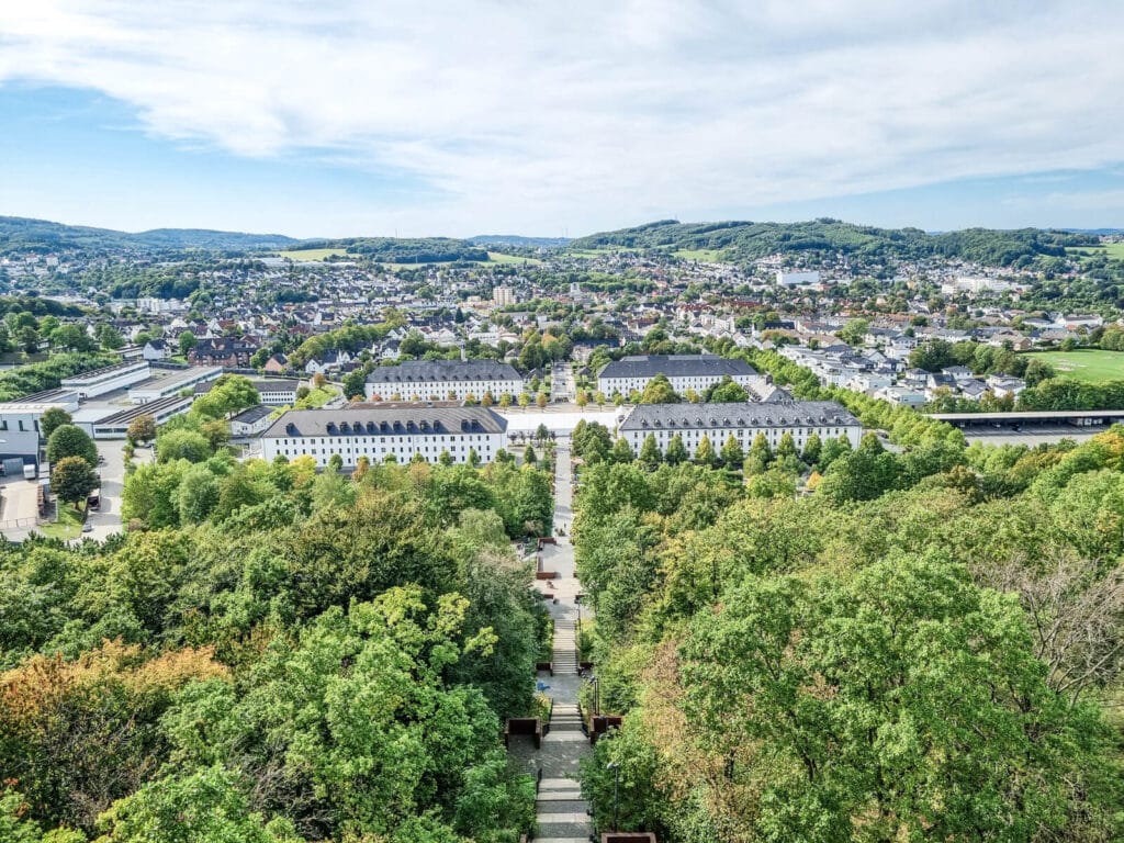 Luftaufnahme einer Stadtlandschaft mit Reihen von weißen Gebäuden, von Bäumen gesäumten Straßen und üppigen grünen Bäumen im Vordergrund, mit Hügeln und verstreuten Häusern im Hintergrund unter einem teilweise bewölkten Himmel.