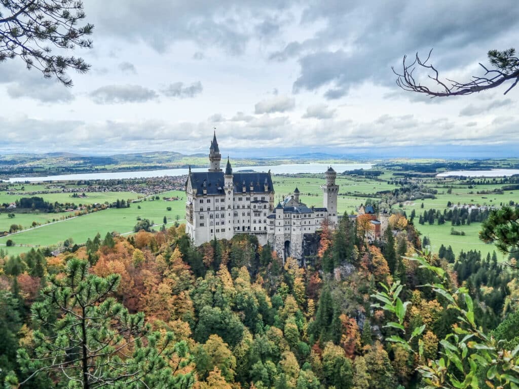 Ein Blick auf Schloss Neuschwanstein, umgeben von bunten Herbstbäumen. Das Schloss steht majestätisch vor einer Kulisse aus sanften grünen Hügeln, entfernten Seen und einem bewölkten Himmel.
