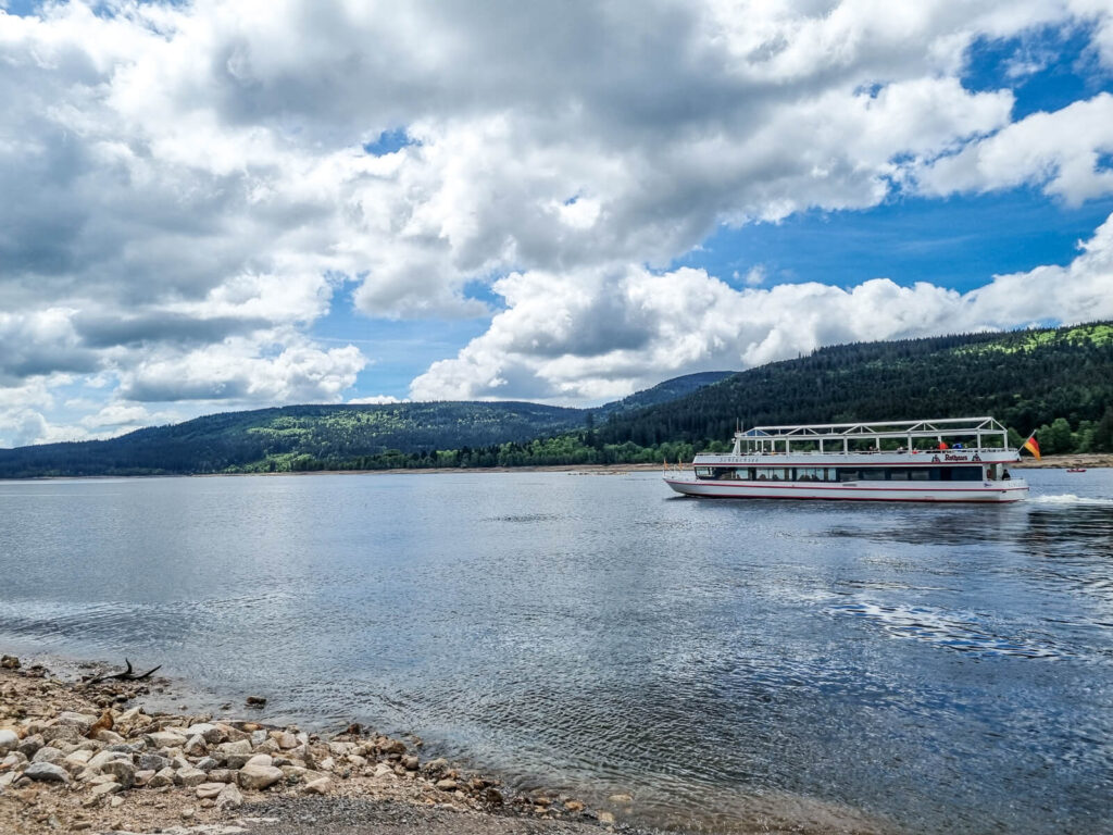 Ein weißes Passagierboot fährt auf einem ruhigen See in der Nähe einer felsigen Küste, mit grünen bewaldeten Hügeln und Bergen im Hintergrund unter einem teilweise bewölkten Himmel.