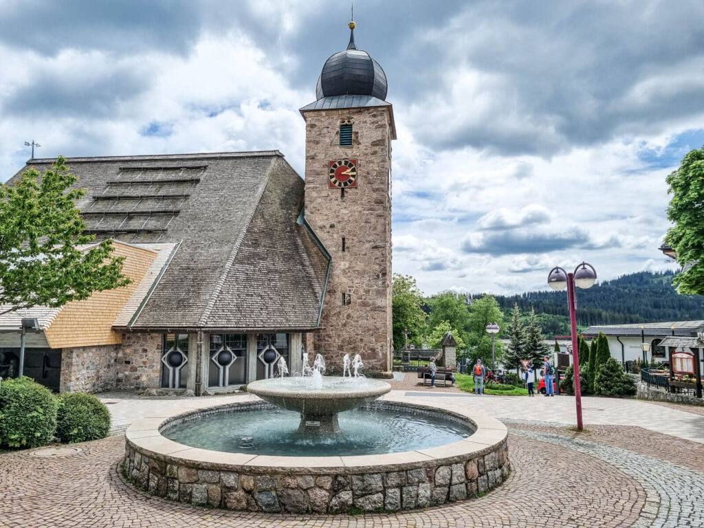 Eine Steinkirche mit einem Glockenturm steht hinter einem runden Brunnen auf einem gepflasterten Platz, umgeben von Bäumen, Laternenpfählen und fernen Hügeln unter einem bewölkten Himmel.