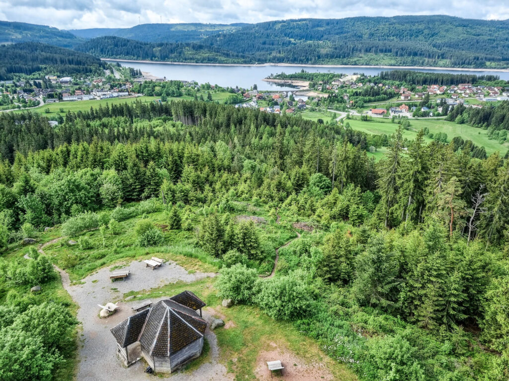 Luftaufnahme eines Waldgebiets mit einem kleinen sechseckigen Gebäude und Bänken im Vordergrund, einem See und einem Dorf im Mittelgrund und baumbedeckten Hügeln im Hintergrund unter einem teilweise bewölkten Himmel.

