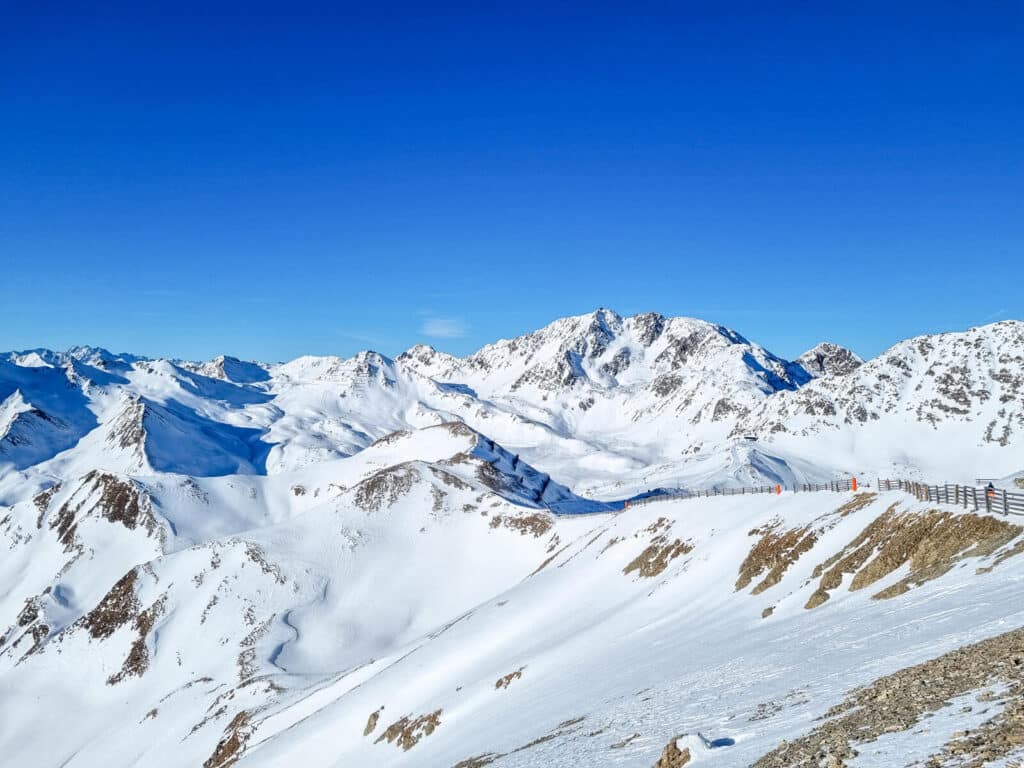 Schneebedeckte Bergkette unter einem klaren blauen Himmel, mit sichtbaren Gipfeln und Tälern. Ein Holzzaun verläuft entlang des Bergkamms und markiert die Grenze. Sonnenlicht hebt den unberührten weißen Schnee hervor und schafft eine helle und ruhige Landschaft.
