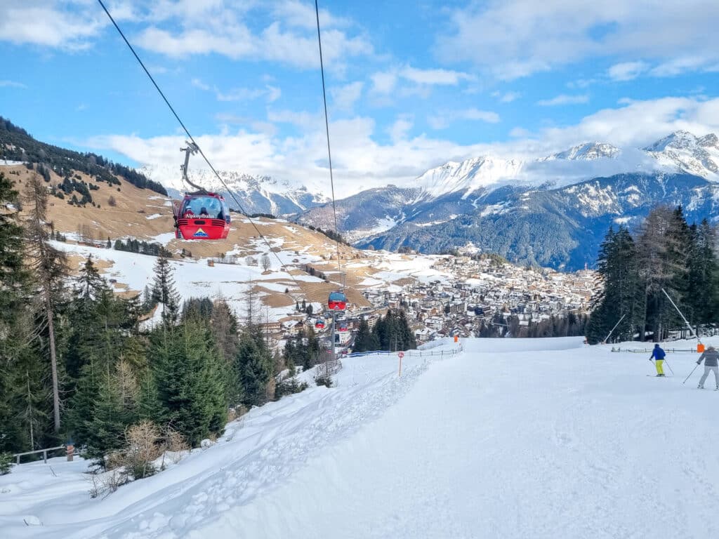 Eine malerische Aussicht auf ein verschneites Skigebiet mit roten Gondeln, die sich darüber bewegen. Menschen fahren auf dem schneebedeckten Hang Ski, umgeben von immergrünen Bäumen. Im Hintergrund erheben sich schneebedeckte Berge unter einem teilweise bewölkten blauen Himmel.
