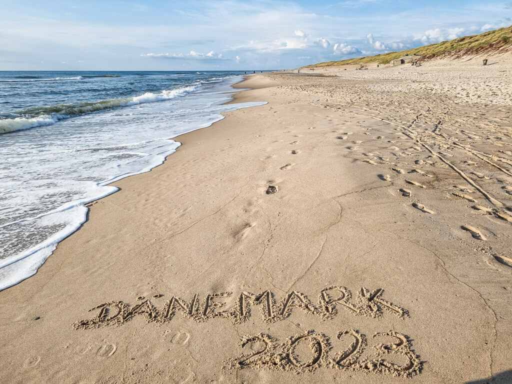 Sandstrand mit einigen Fußspuren und Schriftzug "Dänemark 2023" und das Meer mit Wellen auf der linken Seite
