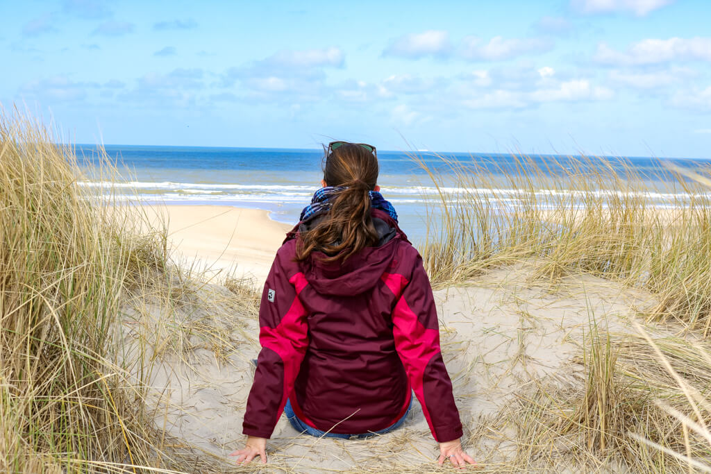 Frau sitzt mit Blick zum Meer auf einer mit Gras bewachsenen Sanddüne