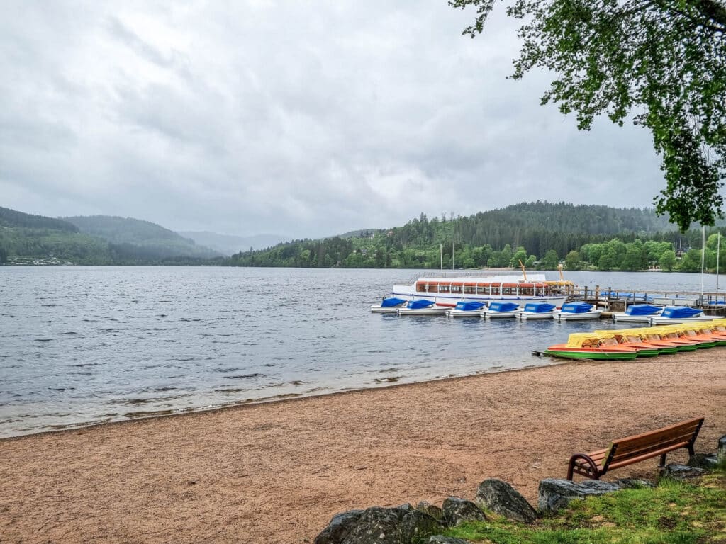 Ein sandiges Seeufer mit einer Holzbank, bunten Paddelbooten, einem angedockten Ausflugsboot und grünen Hügeln und Bäumen im Hintergrund unter einem bewölkten Himmel.