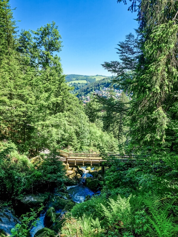 Eine hölzerne Fußgängerbrücke überquert einen Bach, der von üppigen grünen Bäumen und Pflanzen umgeben ist, mit einem Fernblick auf Hügel und ein Dorf unter einem strahlend blauen Himmel.