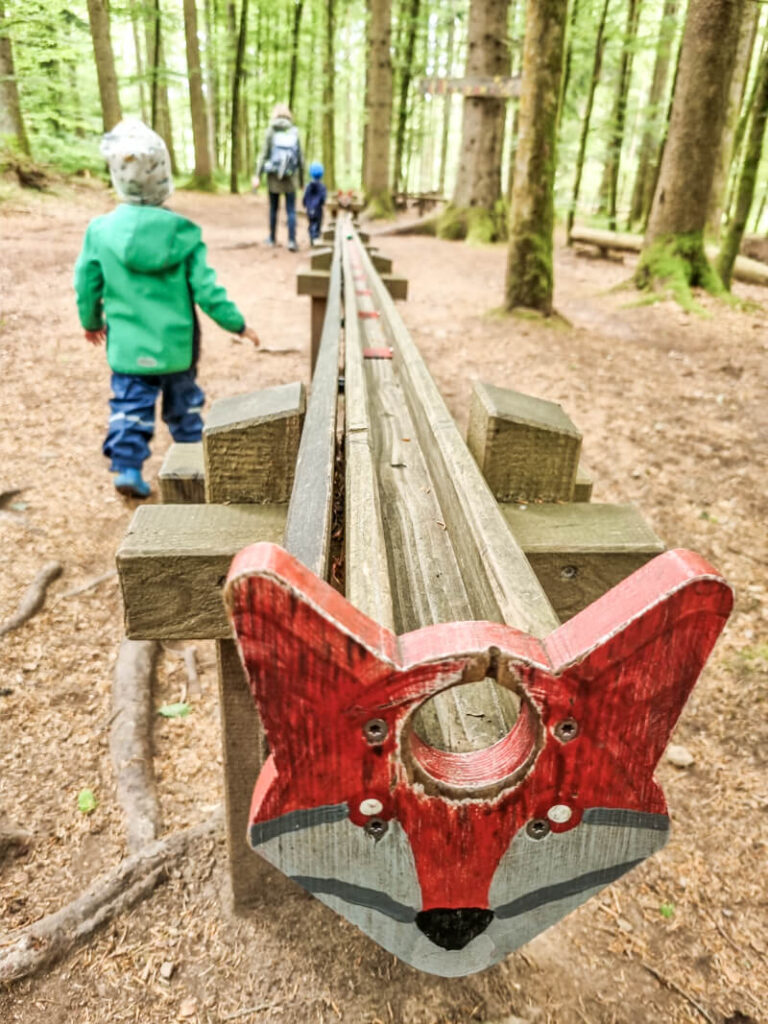 Eine hölzerne Waldkugelbahn mit einem aufgemalten Rotfuchsgesicht am Ende; Kinder und ein Erwachsener (von hinten gesehen) gehen im Hintergrund einen Pfad durch hohe Bäume entlang.