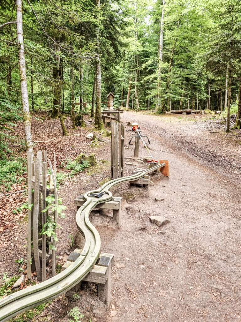 Eine hölzerne Waldkugelbahn mit gewundenen Kanälen steht neben einem Wanderweg in einem üppig grünen Wald. Das Gerät wirkt interaktiv und lädt Kinder zum Spielen und Entdecken ein.

