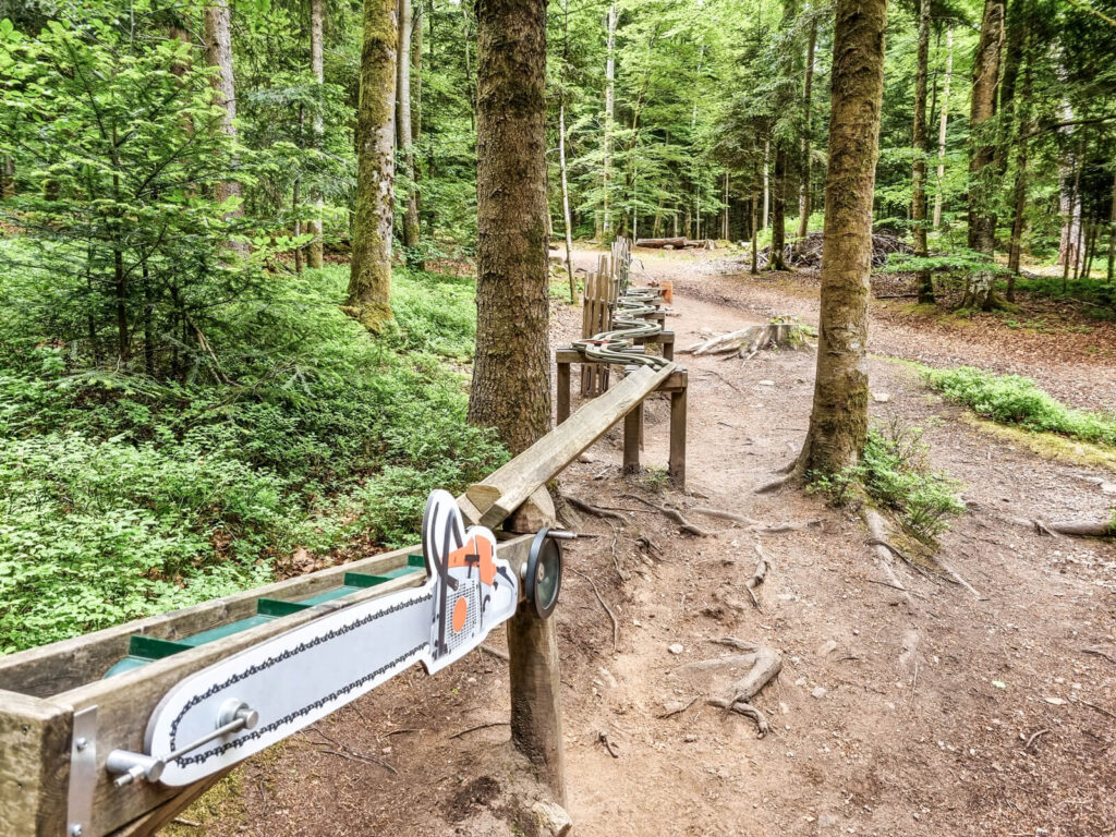 Ein hölzerner Waldpfad bietet interaktive Elemente, darunter eine an einem Geländer befestigte Spielzeug-Kettensäge, umgeben von hohen Bäumen und viel Grün, und ein Feldweg schlängelt sich durch die Szene.