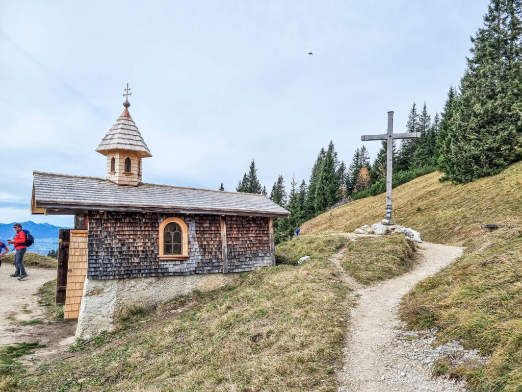 Eine kleine Holzkapelle mit Schindeldach steht auf einem grasbewachsenen Hügel. In der Nähe steht ein großes Holzkreuz. Im Hintergrund sind Kiefern zu sehen, in der Ferne sind Berge zu sehen. Eine Person geht einen Pfad entlang.
