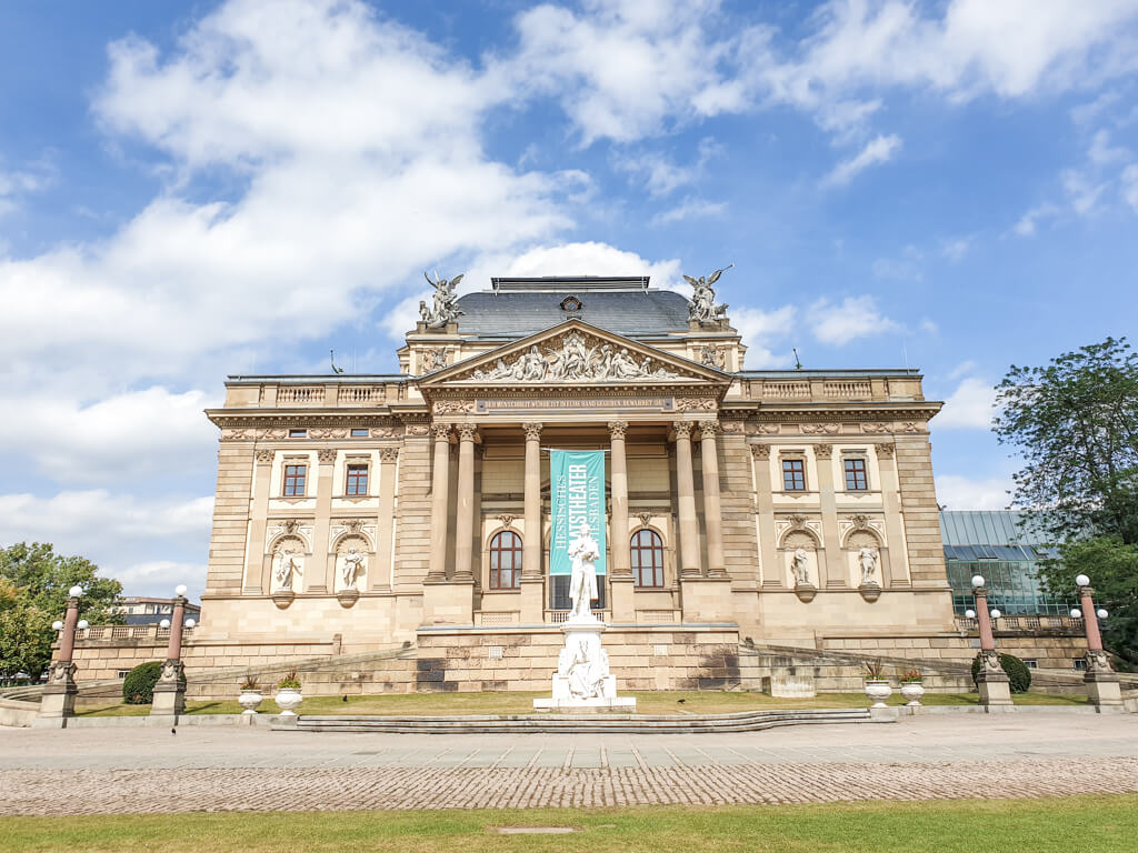 Bild zeigt ein Theater mit einer weißen Statue auf dem Platz vor dem Eingang. Das Gebäude hat sechs Säulen am Eingang mittig, auf beiden Seiten davon nur heller Stein und oben einige Statuen 