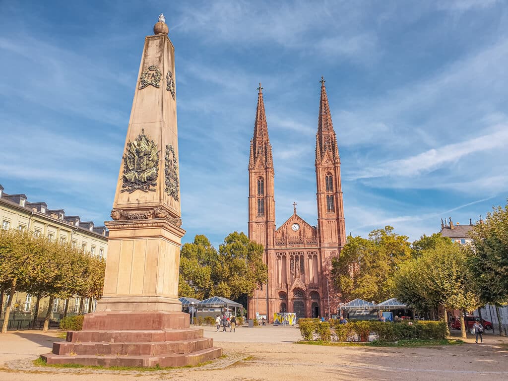 Obelisk aus hellem Stein steht auf einem Platz mit vielen Bäumen, direkt dahinter eine Kirche mit zwei Kirchtürmen