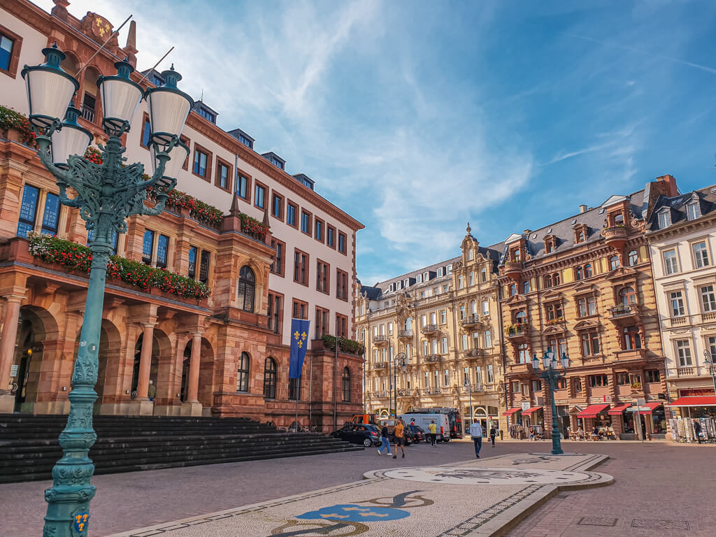 Bild zeigt Marktplatz in Wiesbaden mit historischen Gebäuden, zwei großen Laternen und der Wappeninsel als Mosaik dazwischen