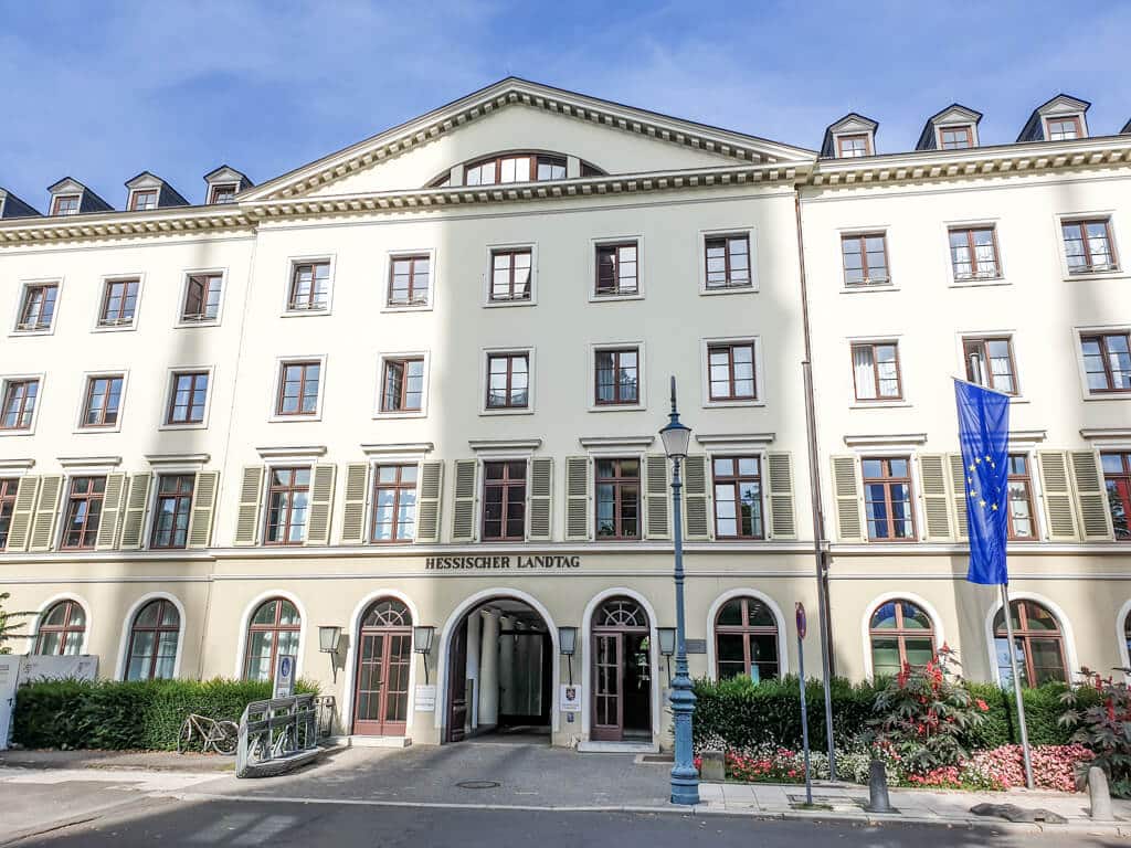 hell gestrichenes Gebäude mit vielen Fensterreihen und runden Fenster- und Torbögen unten; Aufschrift am Gebäude: "Hessischer Landtag"; vor dem Gebäude ist eine Europa-Flagge sichtbar.