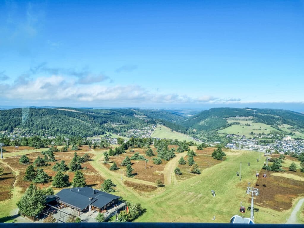Eine malerische Aussicht auf sanfte grüne Hügel mit vereinzelten Bäumen und einigen Gebäuden. Skilifte verlaufen über grasbewachsene Hänge unter einem strahlend blauen Himmel mit vereinzelten Wolken. In der Ferne ist eine kleine Stadt zu sehen.