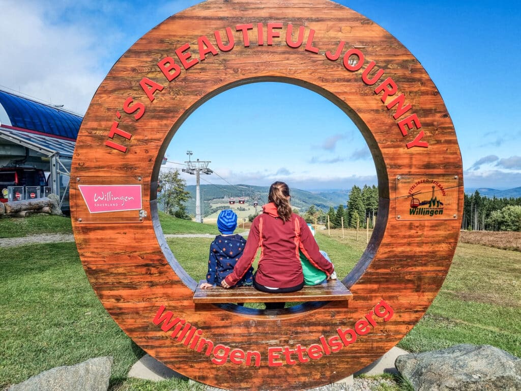 Eine Frau und zwei Kinder sitzen in einem großen runden Holzrahmen mit der Aufschrift "IT'S A BEAUTIFUL JOURNEY" und "Willingen Ettelsberg" und blicken auf eine malerische Berglandschaft.
