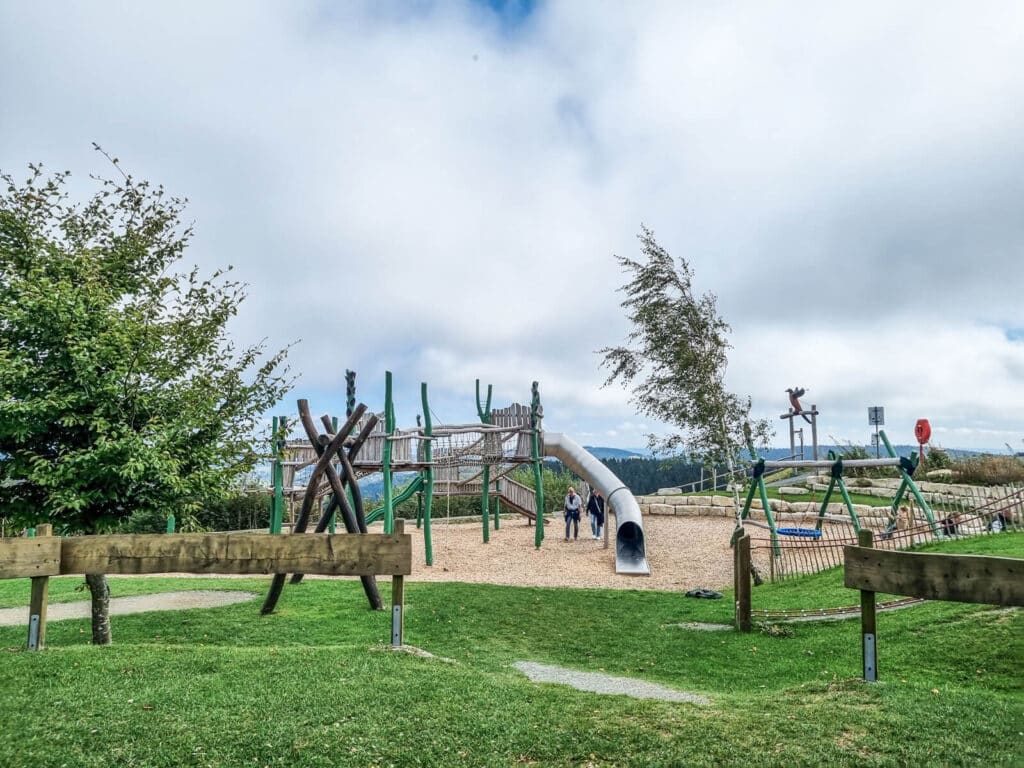 Ein Spielplatz mit Klettergerüsten und einer großen Tunnelrutsche befindet sich auf einer Wiese unter einem bewölkten Himmel. Zwei Personen stehen neben der Rutsche, und Bäume und Holzzäune umgeben den Spielplatz.