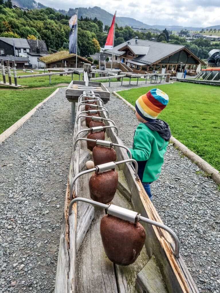Ein kleines Kind mit einer farbenfrohen Mütze steht neben einer Reihe großer Kuhglocken, die auf hölzernen Schienen in einem Park im Freien montiert sind, mit grünem Gras, Gebäuden und Hügeln im Hintergrund.


