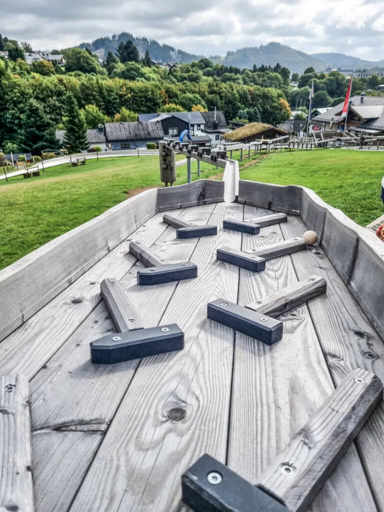 Nahaufnahme eines hölzernen Tischspiels im Freien, mit schwarzen Hindernissen und einem Ball, auf grünem Gras mit Häusern, Bäumen und Hügeln im Hintergrund unter einem bewölkten Himmel.