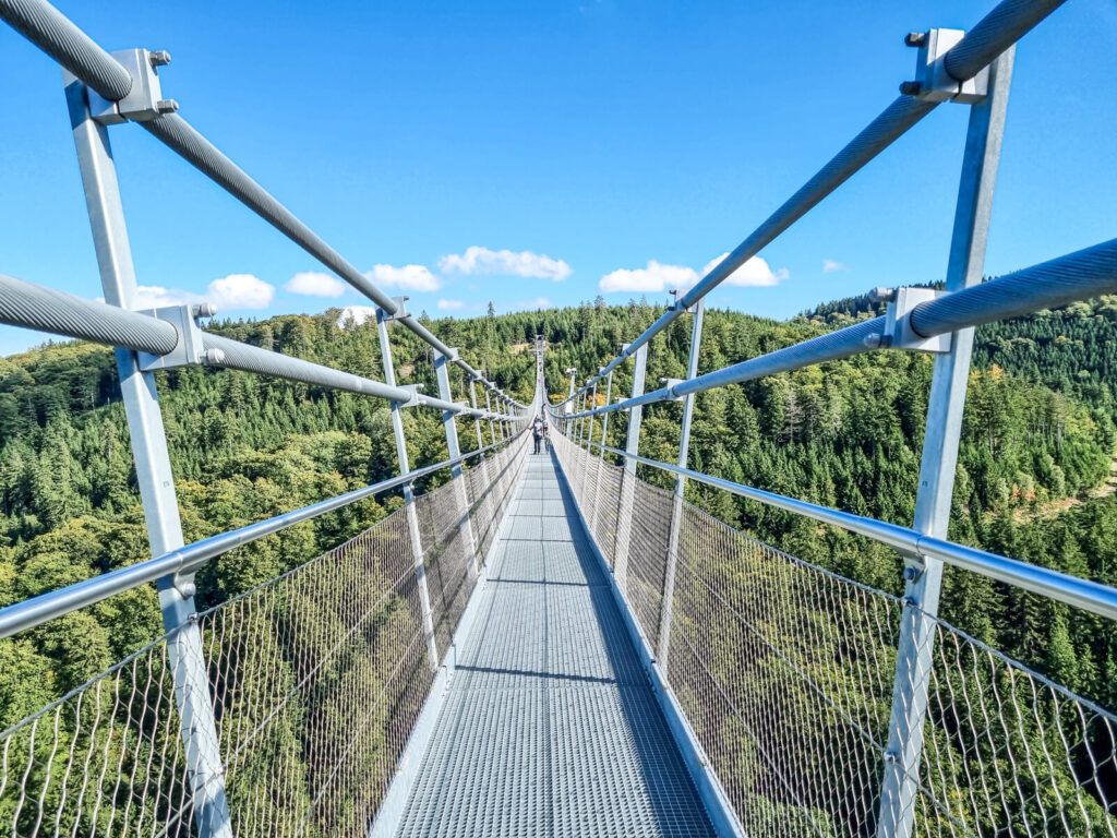 Eine lange Hängebrücke mit Metallgeländer führt über einen üppigen grünen Wald und zu fernen Hügeln unter einem strahlend blauen Himmel mit vereinzelten Wolken.