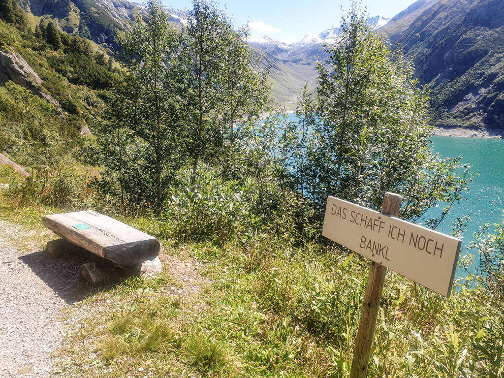 kleine Holzbank mit Schild "Das Schaff ich noch Bankl" im Grünen, dahinter ein blauer Bergsee